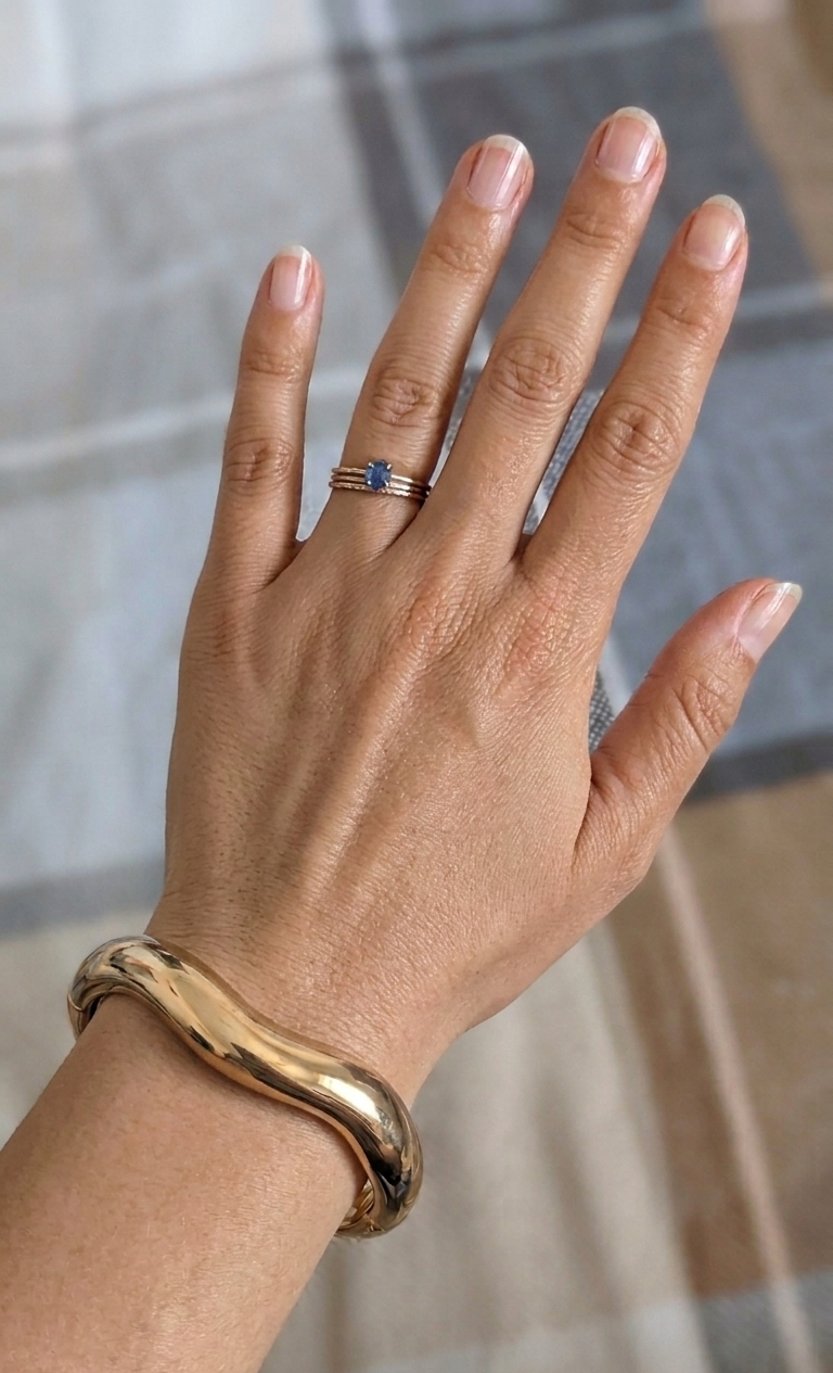Hand wearing a gold bracelet and ring with a blue stone on a neutral background
